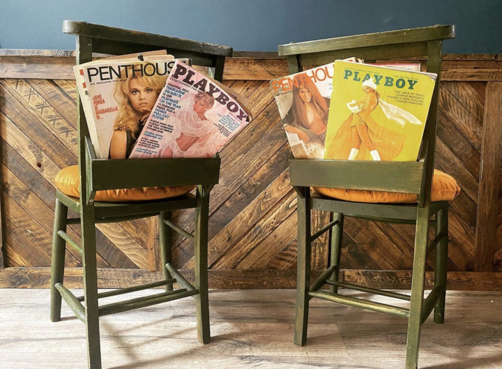 Two green, up-cycled church chairs side by side with vintage Playboy magazines in the bible holders. The back drop is a teal blue wall with panelling on the lower half made from rustic scrap wood.