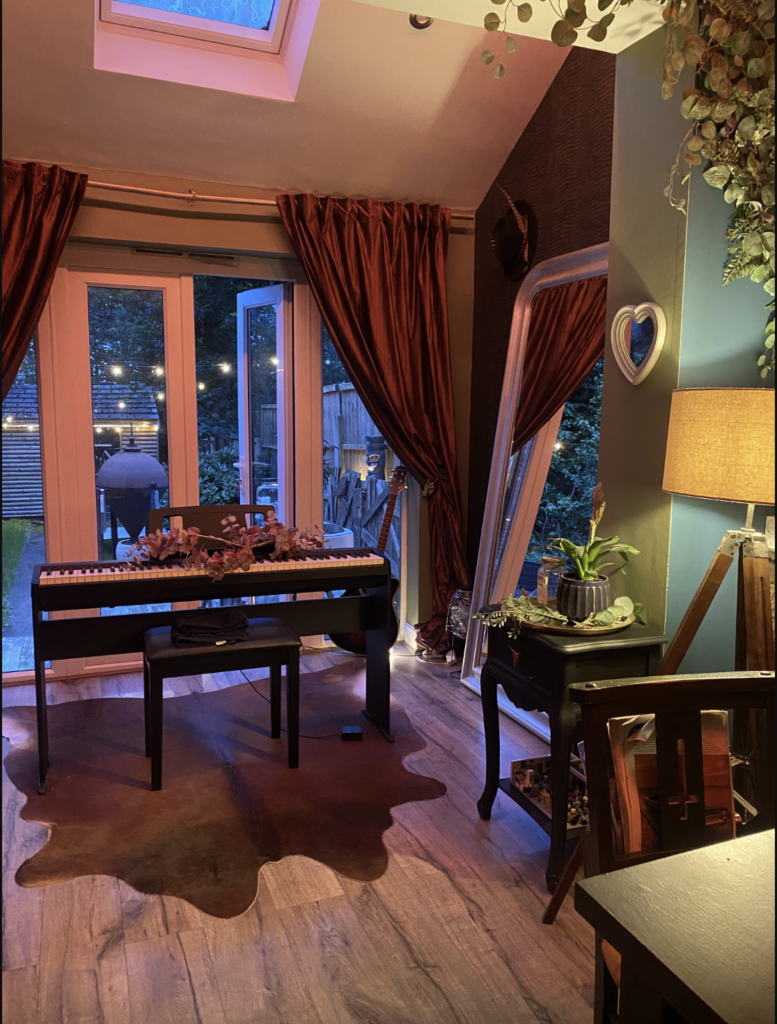 An electric piano sits atop a deer hide rug in a cosy corner of the ground floor. Heavy, copper coloured velvet curtains adorn the wide patio windows that look out to a garden lit up with string lights.