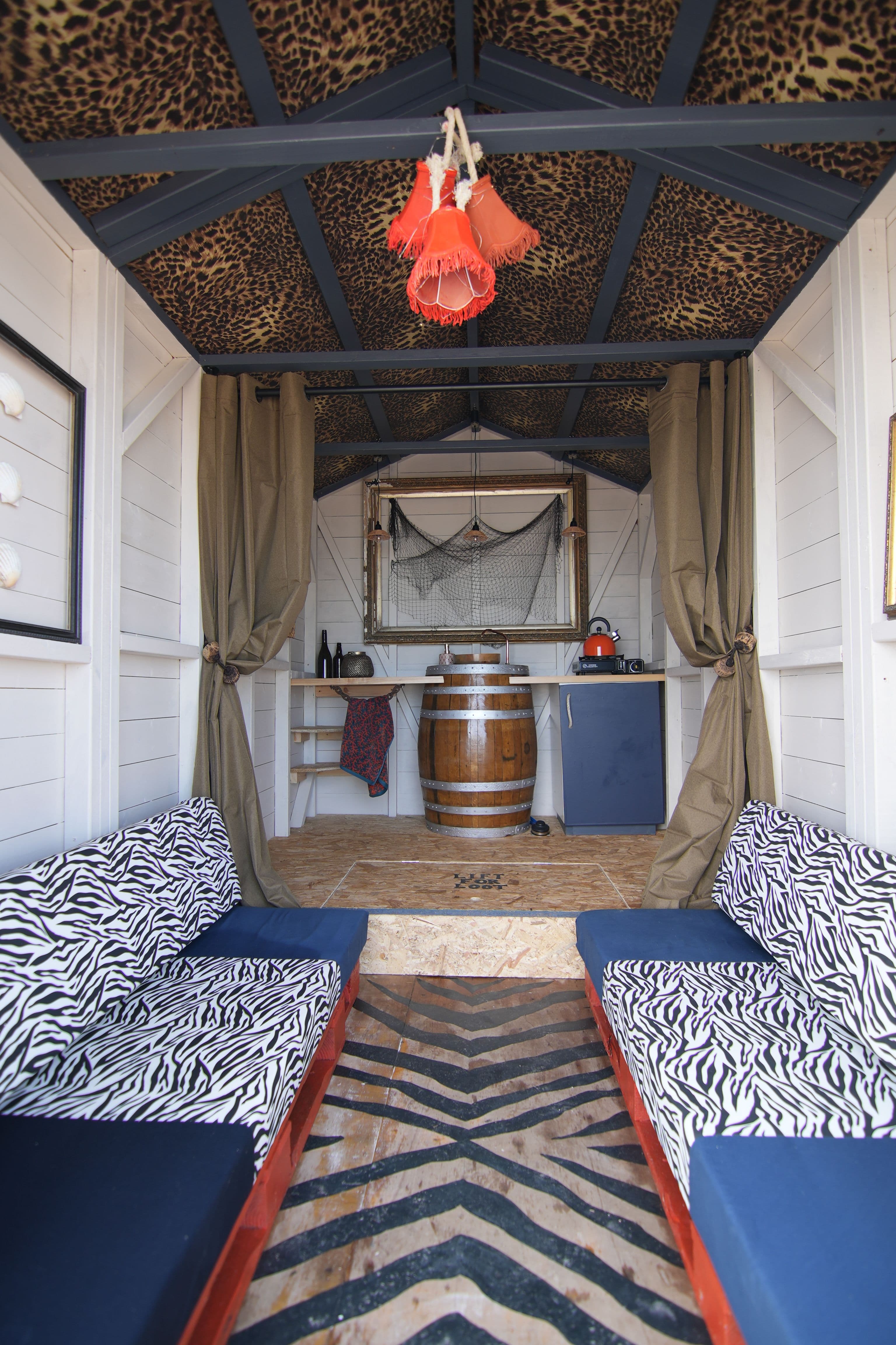 A rock n roll beach hut interior, with zebra print hand painted floor, leopard print wallpaper on the ceiling, a conversation pit and a barrel sink on a platform at the back.