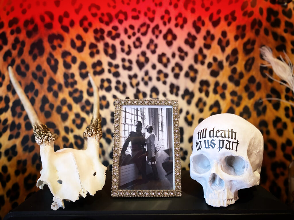 Leopard print wallpaper backdrop with a deer skull cap, framed wedding photo and a replica human skull in front of it. maximalist interiors